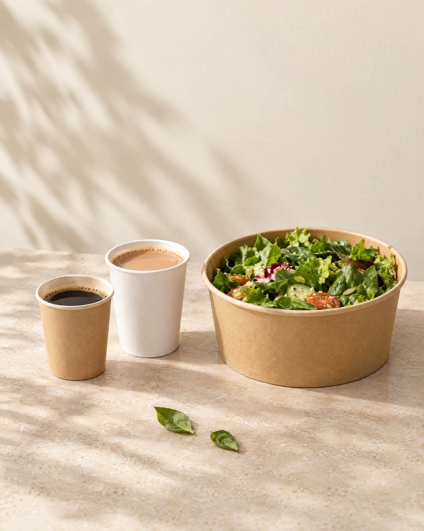 A small kraft paper cup of black coffee and a small white paper cup of chai beside a kraft paper bowl of green salad, on a sand-coloured stone surface.